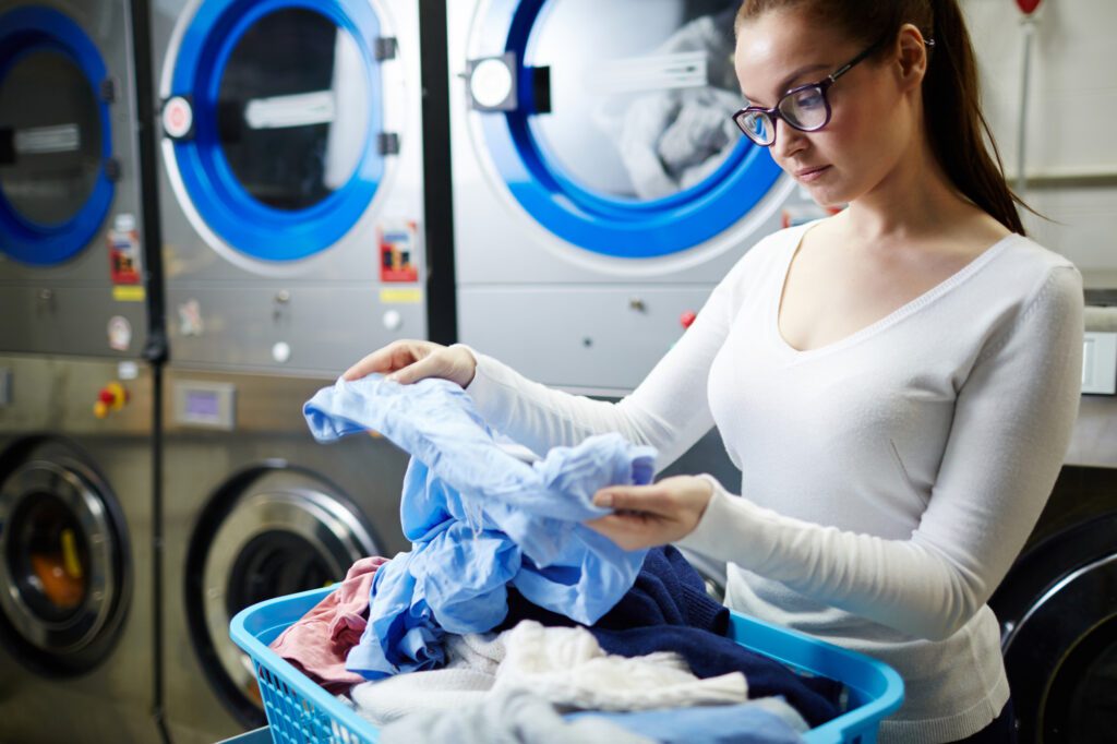 A woman in glasses examines clothes from a laundry basket in front of industrial washing machines at a laundromat.