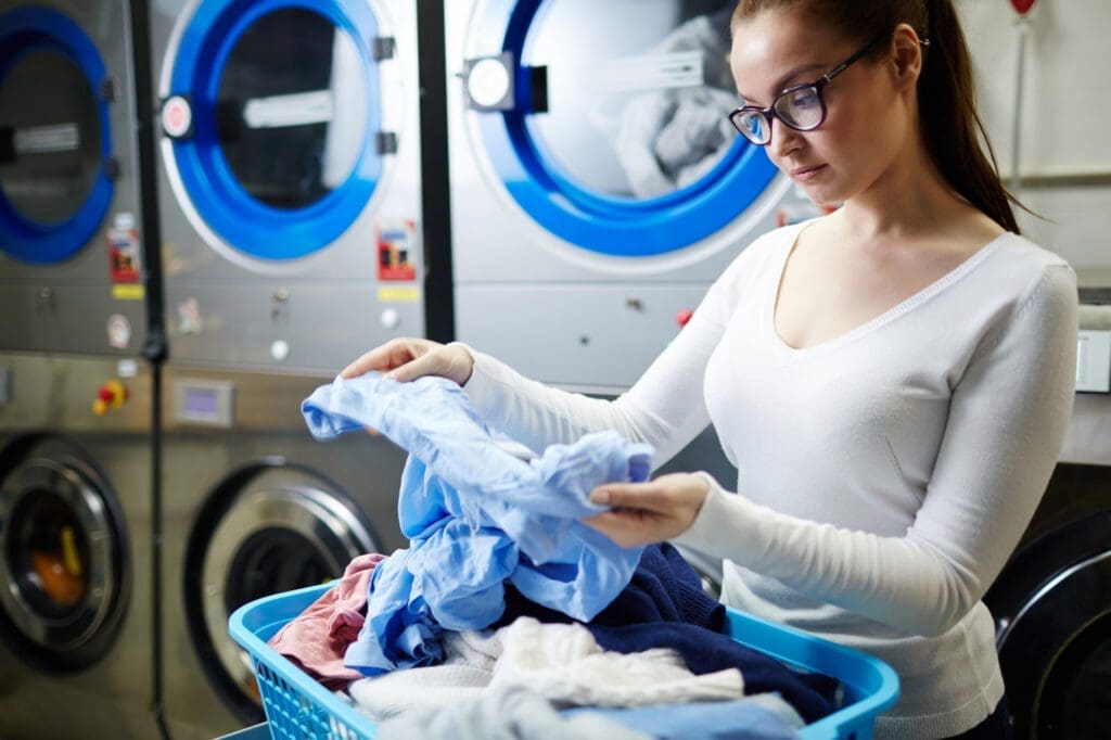 A woman in glasses examines clothes from a laundry basket in front of industrial washing machines at a laundromat.