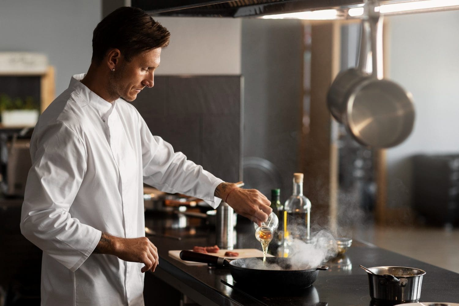 A chef in a white coat pours liquid into a steaming pan on a stovetop in a modern kitchen.