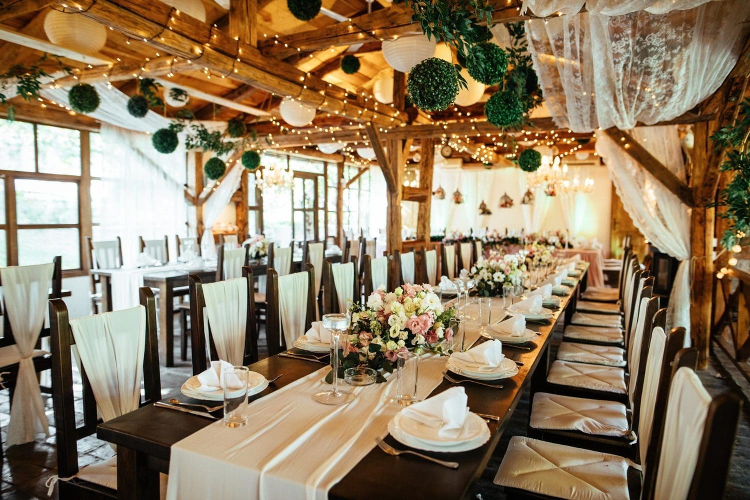 Long banquet tables are set for a formal event in a rustic, wooden venue decorated with string lights, white drapes, greenery, and floral centerpieces.