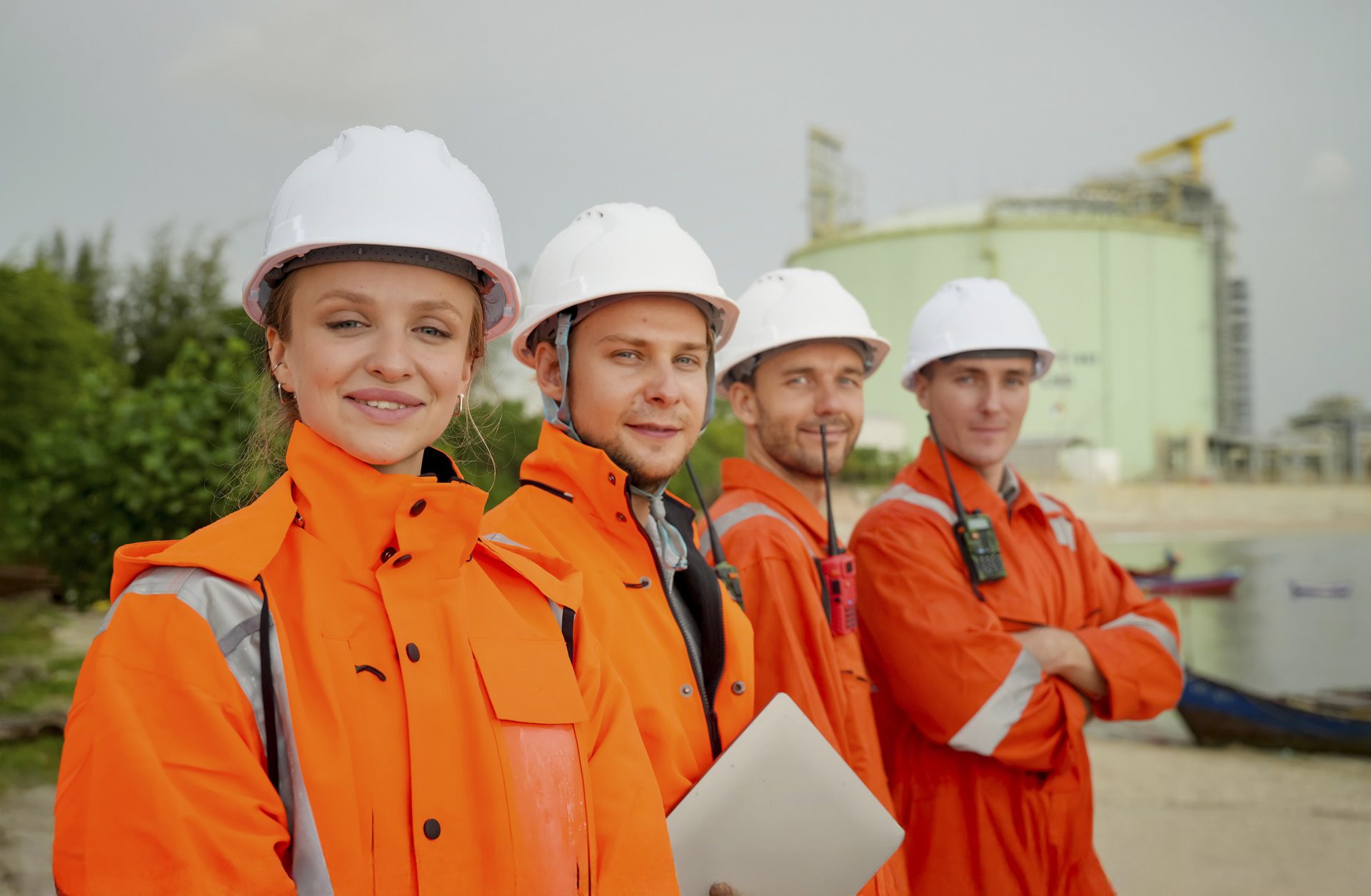 Four workers in orange safety jackets and white hard hats stand outdoors at an industrial site, with a large storage tank visible in the background.