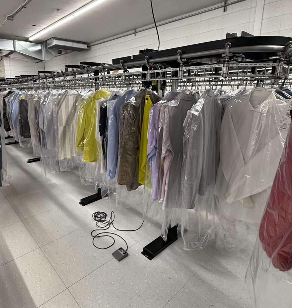 Clothes covered in plastic hang on a conveyor rack in a dry cleaning facility. A power cord and foot pedal lie on the tiled floor below.