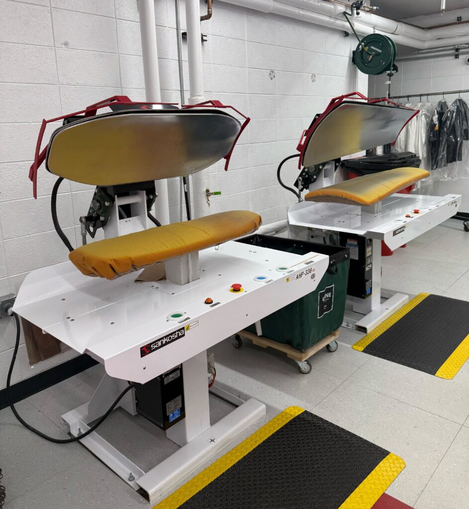 Two industrial steam press machines with padded boards are situated in a clean laundry facility, used for pressing clothes.