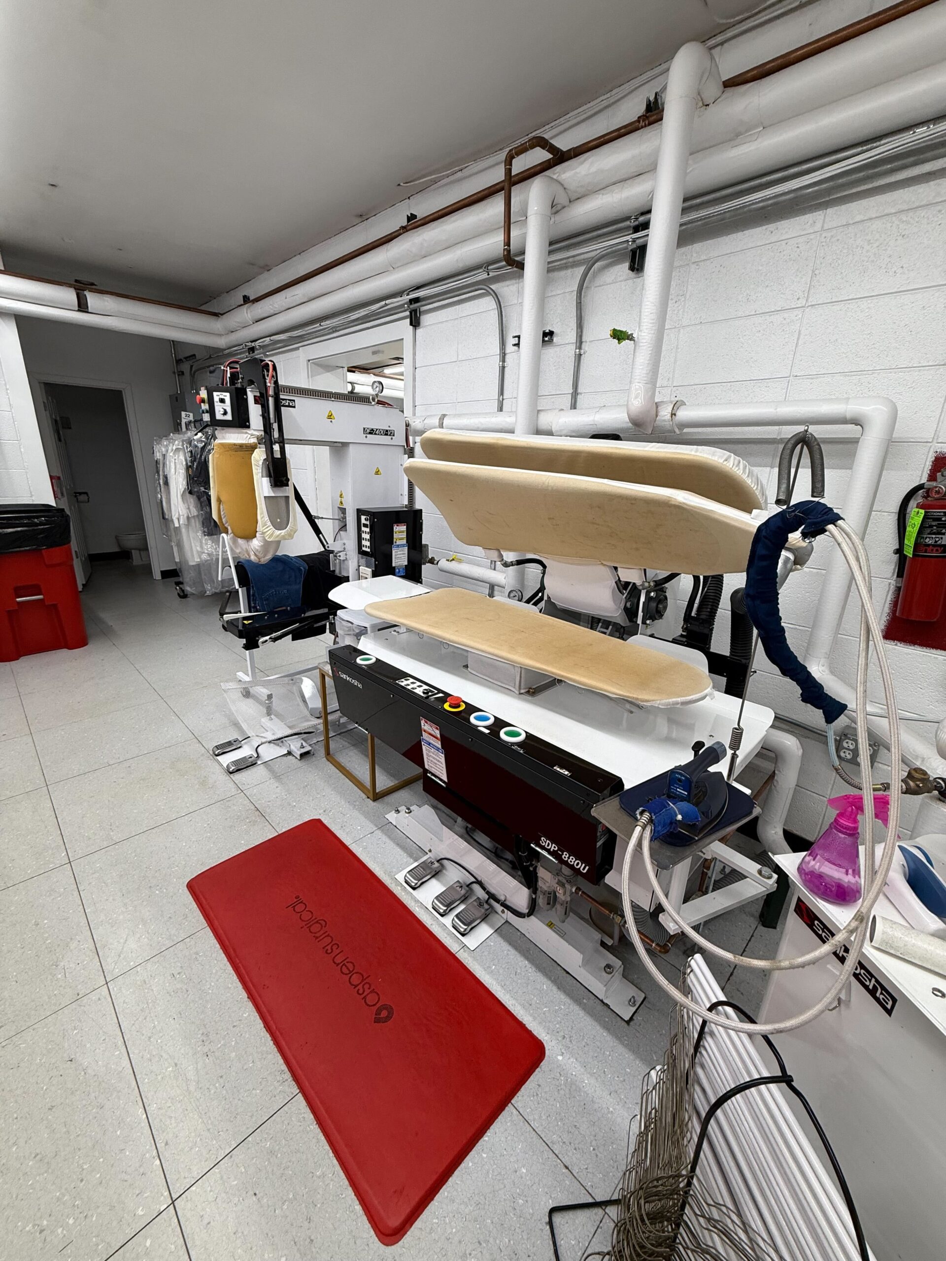 A commercial laundry press with padded plates and control buttons stands in a utility room, surrounded by pipes, hangers, and cleaning equipment.