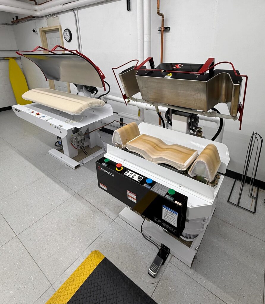 Two industrial laundry pressing machines with padded surfaces and control panels are positioned in a clean, tiled room with white walls.