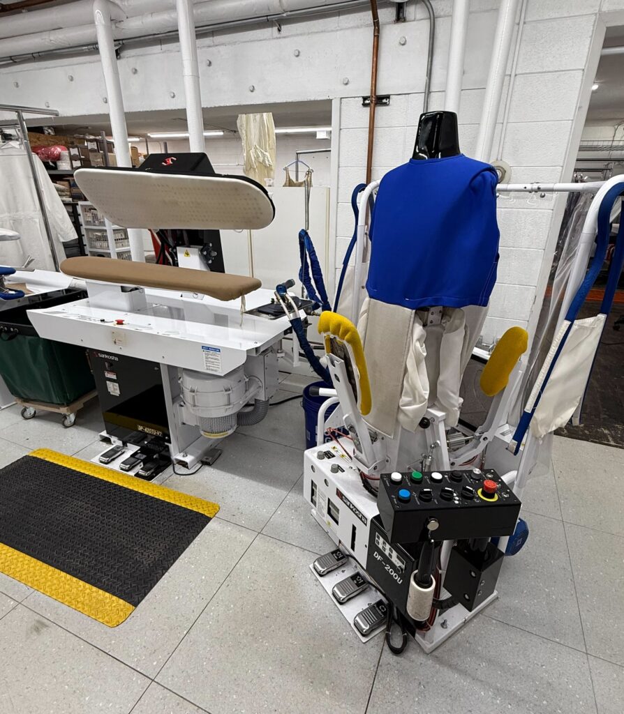 A commercial garment pressing machine and a shirt-finishing machine with a blue cover are positioned in a laundry or dry cleaning facility.